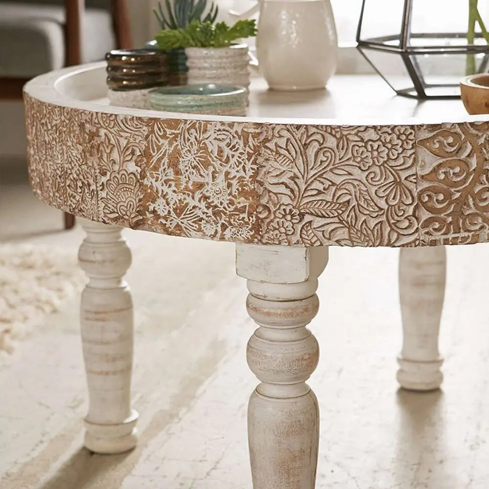 Round wooden table with intricate carvings and white legs in a room setting.