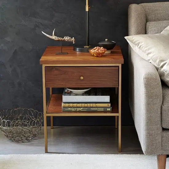 An Alberto end table with a natural wood finish and metal base, placed in a room beside a sofa, with books and a decorative item on top.