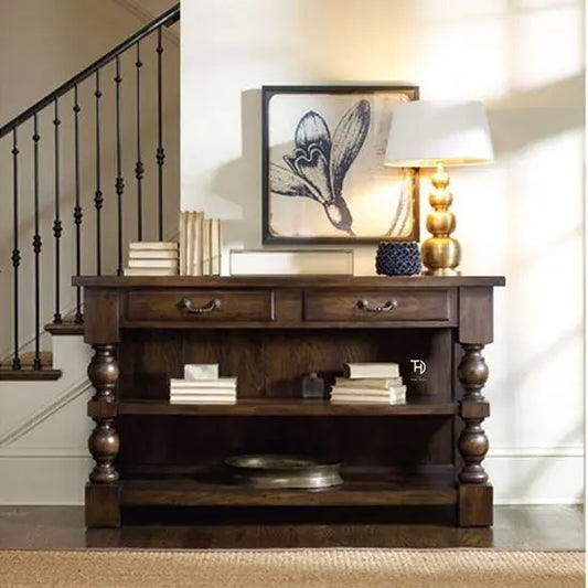 A wooden Shiva TV console with two drawers, a shelf, and a walnut finish, placed in a room with decorative items on top and a staircase in the background.