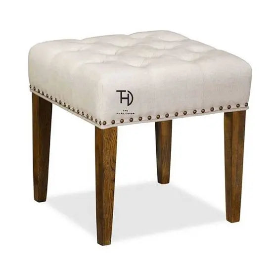 An upholstered stool with a tufted top and head nail detailing, featuring four wooden legs in a natural finish.