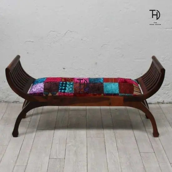 A two-seater bench with a curved design, featuring a multicolored patchwork fabric seat and a wooden structure with a honey finish.