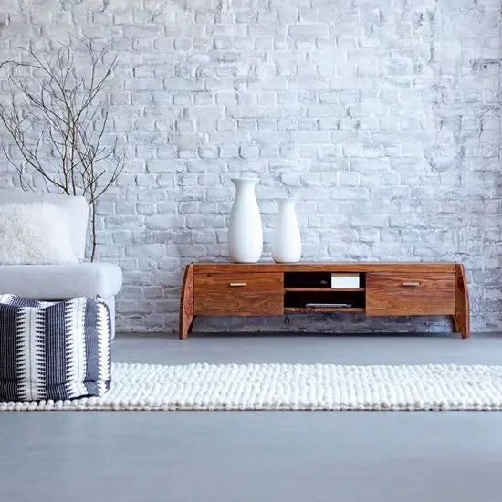 A solid sheesham wood Rhino TV cabinet with two wooden doors and open shelves, placed against a brick wall. The cabinet is brown in color, with natural wood grain visible.