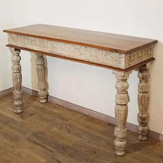 A rustic white console table with intricate hand carving on the side and legs, with a natural wood top.