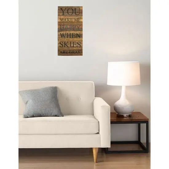 A piece of wall art hanging above a sofa, featuring text, with a lamp and wooden side table in the background.