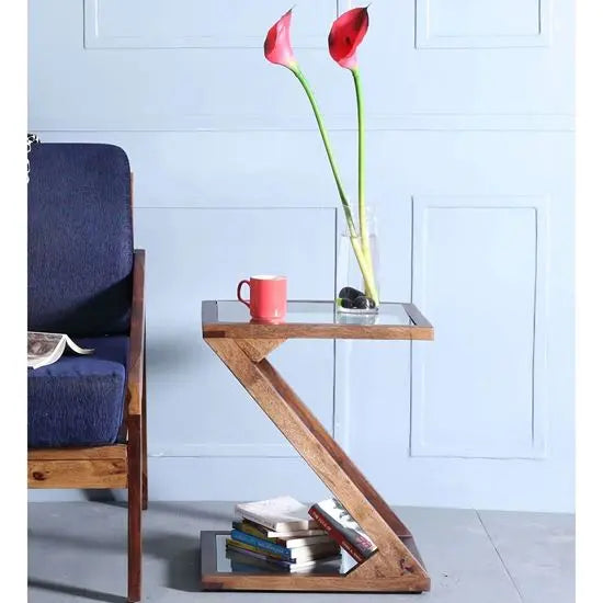 A Z-shaped end table with a glass top placed on it, with sheesham wood frame.