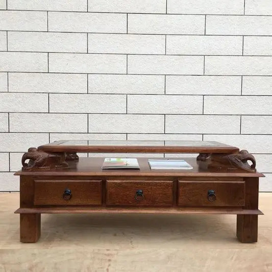 A rectangular brown Asgil coffee table made of mango wood with three drawers and carved elephant details on the sides.