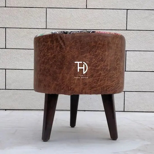 A round brown leather pouf with a patchwork fabric top and three wooden legs.