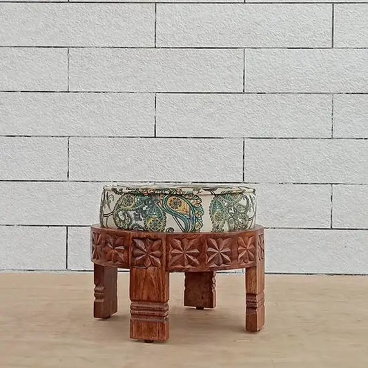 A hand-carved wooden footstool with a round shape and a multicolored velvet fabric on top, intended for indoor use.