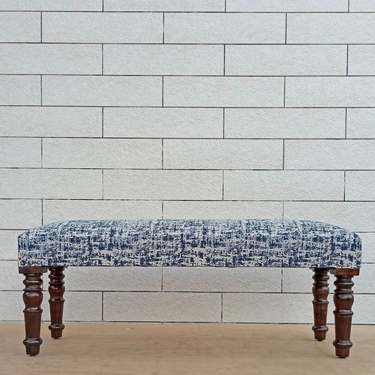 A bench with a upholstered blue and white patterned seat and wooden legs, placed against a brick wall.