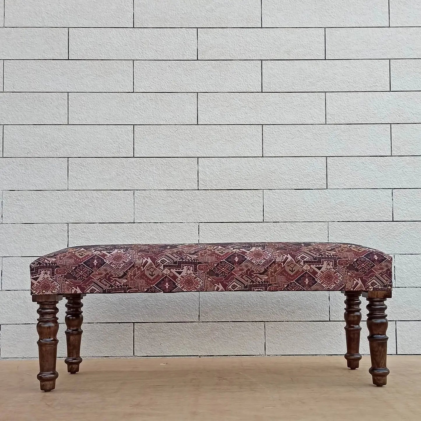 A rectangular bench rug with a geometric pattern, displayed on a bench with wooden legs against a white brick background.