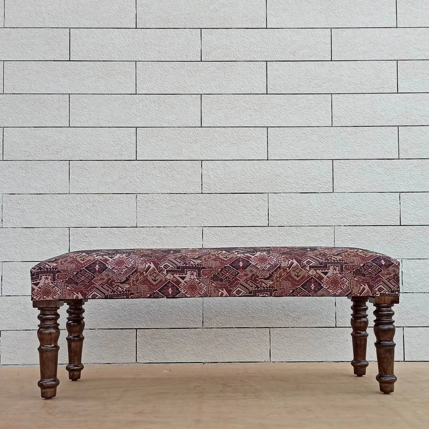 A rectangular bench rug with a geometric pattern, displayed on a bench with wooden legs against a white brick background.