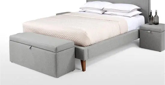 A gray upholstered storage bench at the foot of a bed, with a closed lid and a cushion on top for seating.