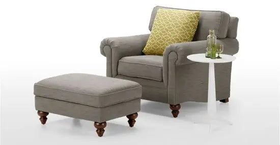 A brown Della Storage Footstool with turned-round legs, placed in front of an armchair.