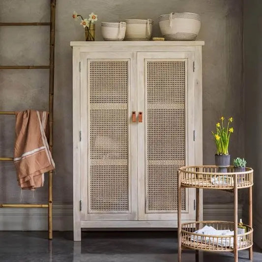 A white wash finish cane almira with two doors, placed in a room with various decor items around it.