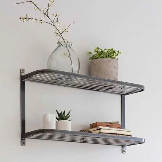 A metal wall-mounted shelf with two levels, displaying a glass vase with flowers, a beige pot plant, a white vase, a small wooden bookshelf, and a few books.
