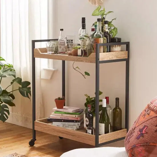 A serving trolley made of mango wood with metal legs, featuring a shelf with bottles and a lower shelf with a potted plant and books, placed in a home setting.
