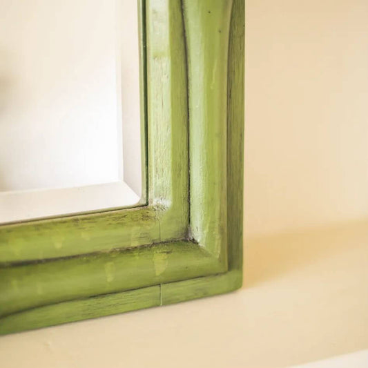 A close-up of a distressed green wooden picture frame corner, showing the painted wood grain and a hint of the white mirror glass inside.