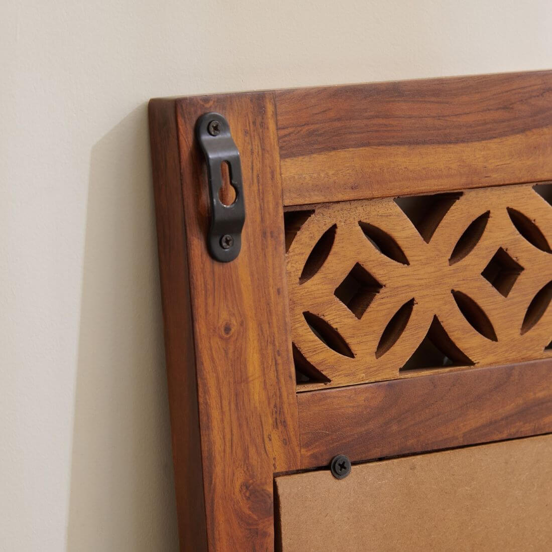 Close-up of a wooden wall-mounted cabinet or shelf corner with a carved geometric cutout panel, dark metal wall bracket, and a light wood backing board wall.