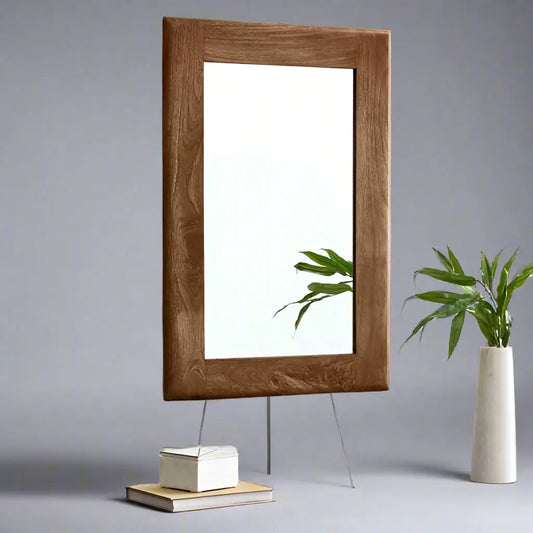 A rectangular mirror with a wooden frame, standing on a grey background with a plant to the side.