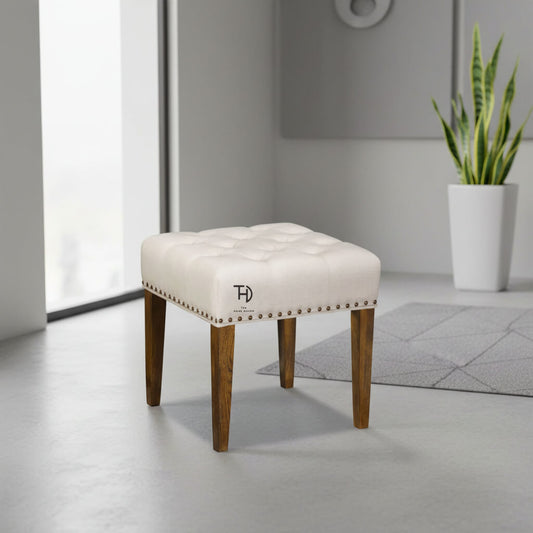 An upholstered stool with a tufted top and head nail detailing, featuring four wooden legs in a natural finish.