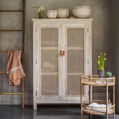 A white wash finish cane almira with two doors, placed in a room with various decor items around it.