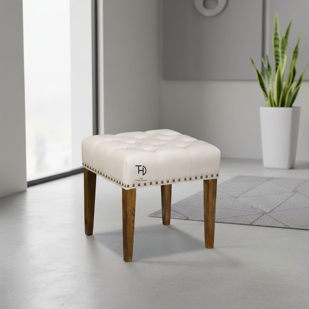 An upholstered stool with a tufted top and head nail detailing, featuring four wooden legs in a natural finish.