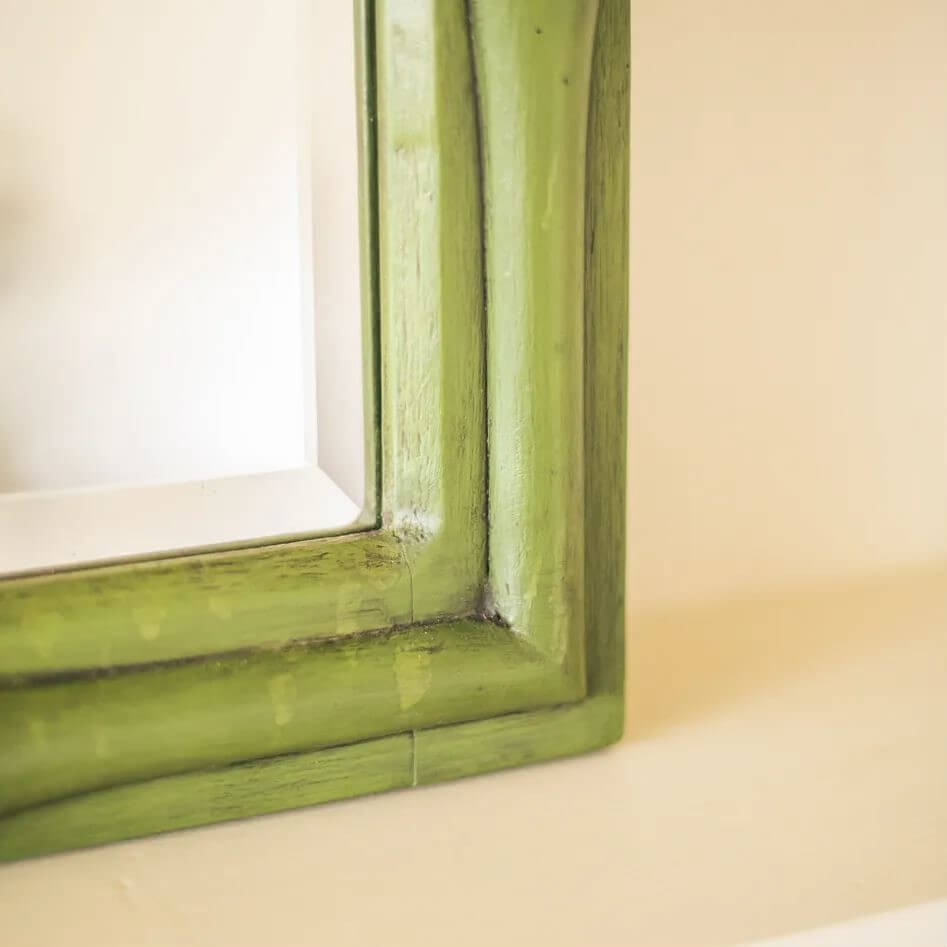 A close-up of a distressed green wooden picture frame corner, showing the painted wood grain and a hint of the white mirror glass inside.