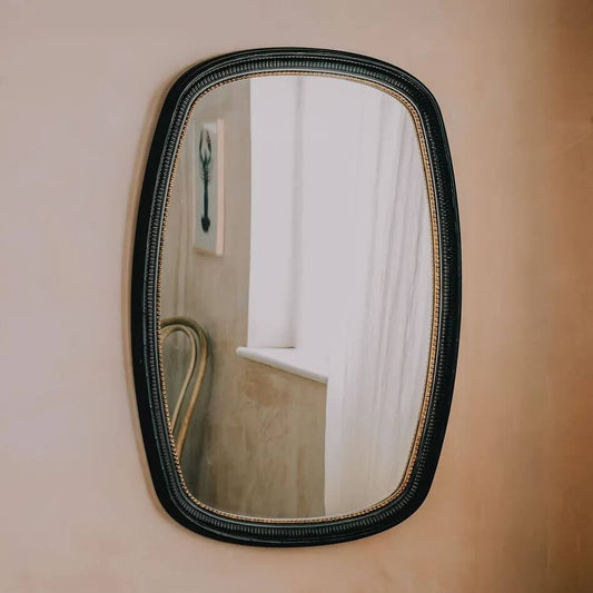 An oval wall mirror with a textured black frame and a gold inner trim.