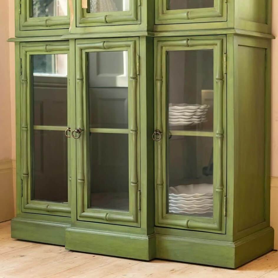 An olive green painted wooden cabinet with glass-paneled doors and brass ring handles.