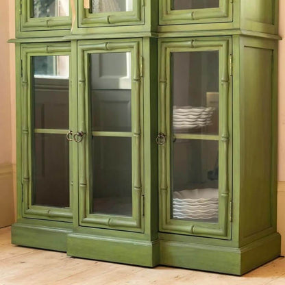An olive green painted wooden cabinet with glass-paneled doors and brass ring handles.