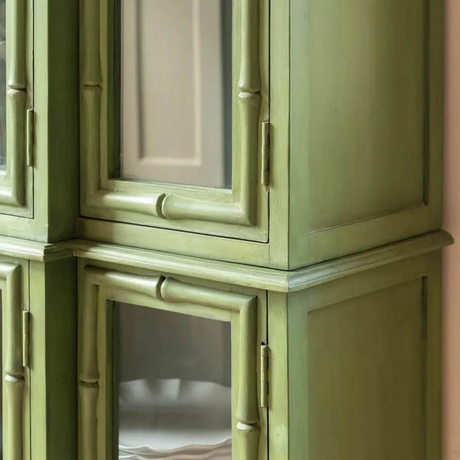 A close-up of a painted wooden cabinet with a distressed sage green finish and bamboo-style trim around the glass-front doors.