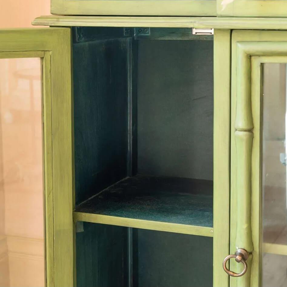 A close-up of a green wooden cabinet with glass doors and a brass ring pull, showing an empty interior shelf.