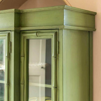 A close-up of a painted wooden cabinet corner with a sage green finish and glass-front doors.