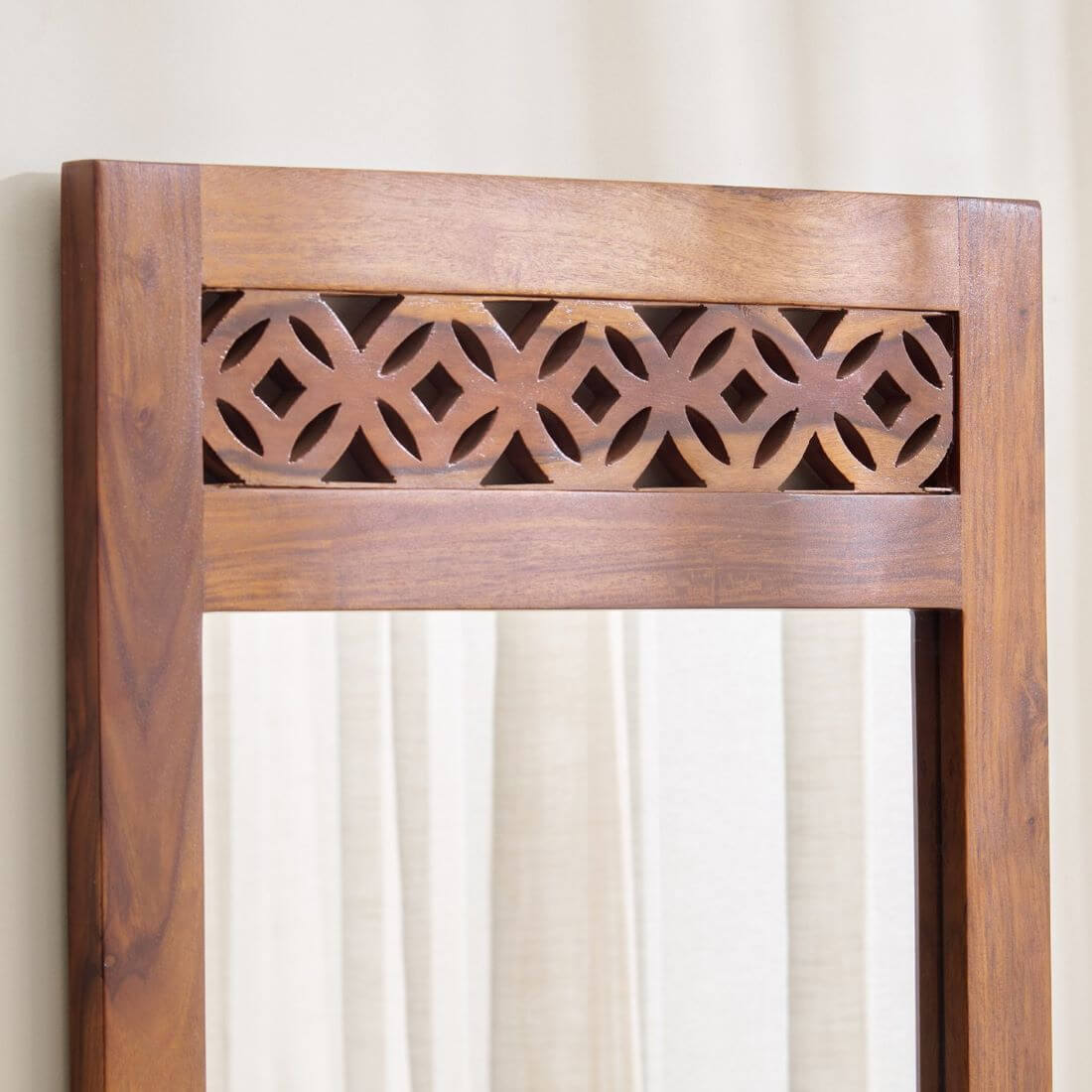A close-up of a wooden mirror frame with a warm medium-brown finish and an ornate geometric cutout pattern across the top.