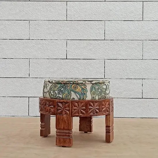A hand-carved wooden footstool with a round shape and a multicolored velvet fabric on top, intended for indoor use.
