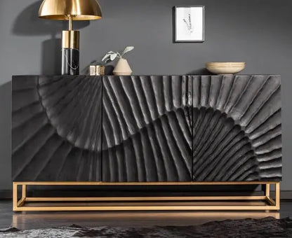 A black mango wood sideboard with three doors and a metal base, featuring a wave-like pattern on the doors, placed in a room with a gold lamp and decorative items.