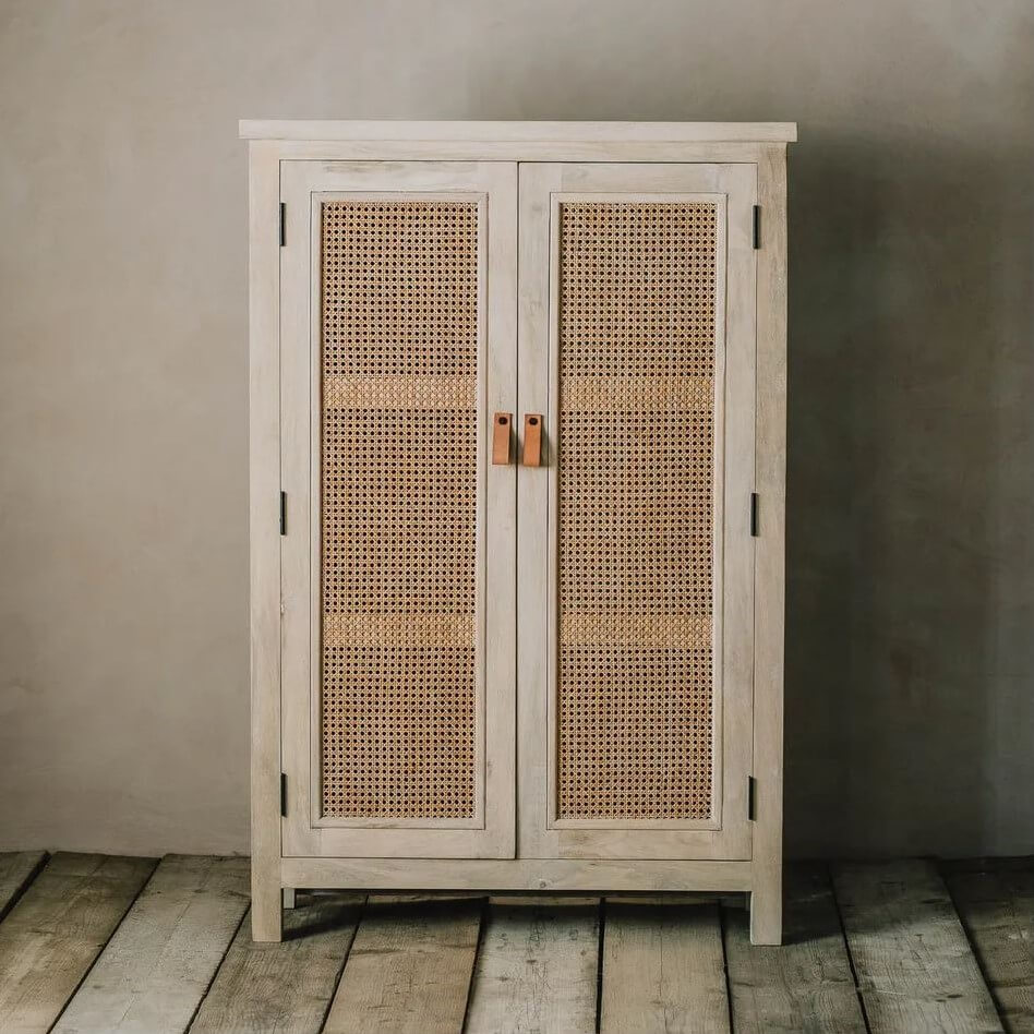 A rustic two-door cabinet with woven cane panels on the doors, finished in a light whitewashed wood tone with dark metal hinges and small wooden pull handles.