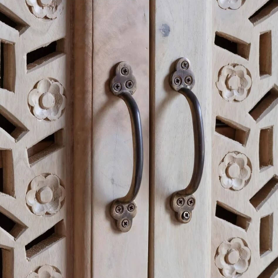 Close-up of two dark metal cabinet handles mounted on a light natural wood door with carved geometric and floral patterns.