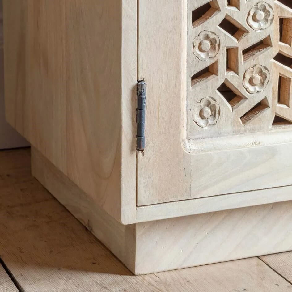 Close-up of a light natural wood cabinet corner with a carved geometric floral door panel and a dark metal hinge.