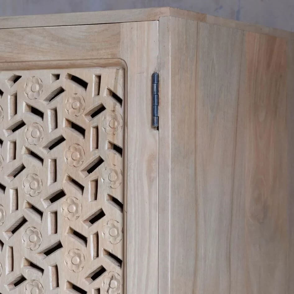 Close-up of a light natural wood cabinet corner showing a carved geometric patterned door panel, a dark metal hinge, and the side wood grain.